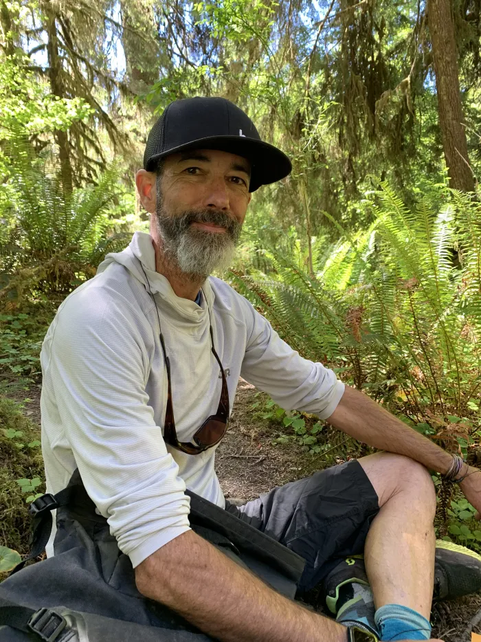 Portrait of Brian Miesch, bearded and smiling in a cap, sitting among ferns.