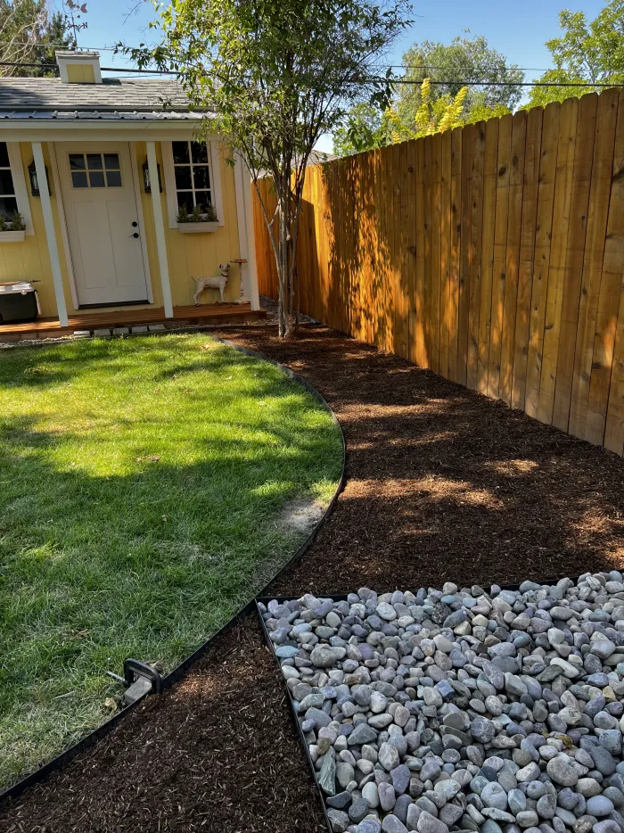 Side yard with a curved lawn edge, a strip of river rock and mulch, and a tall cedar fence.