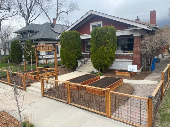 Front yard garden with raised beds and an arbor inside a redwood and wire fence, in front of a brick bungalow.