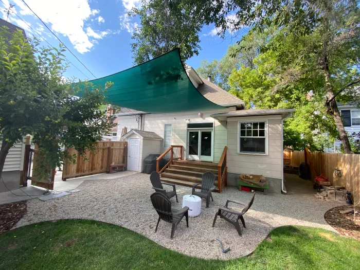Backyard gravel patio with Adirondack chairs under a green shade sail, edged by a curving lawn.