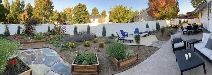Panorama of a backyard with flagstone paths, raised garden beds, drought tolerant planting, a dining set and lounge chairs along a white fence.
