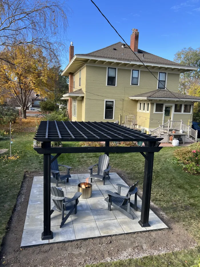 Black pergola over a square paver patio with a fire pit and chairs, beside a yellow two-story house.