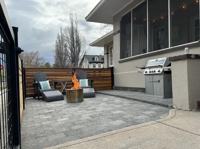 Paver patio with a round fire pit, two Adirondack chairs, a built-in grill and a horizontal cedar screen.