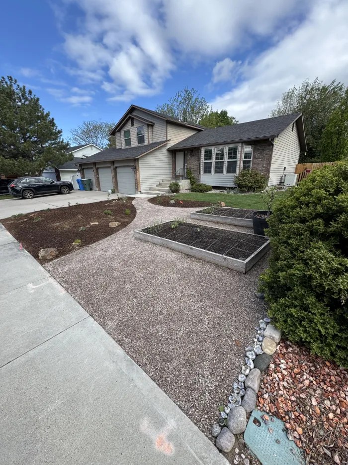 Front yard of gravel and flagstone with raised planting beds and river rock, beside a house and sidewalk.